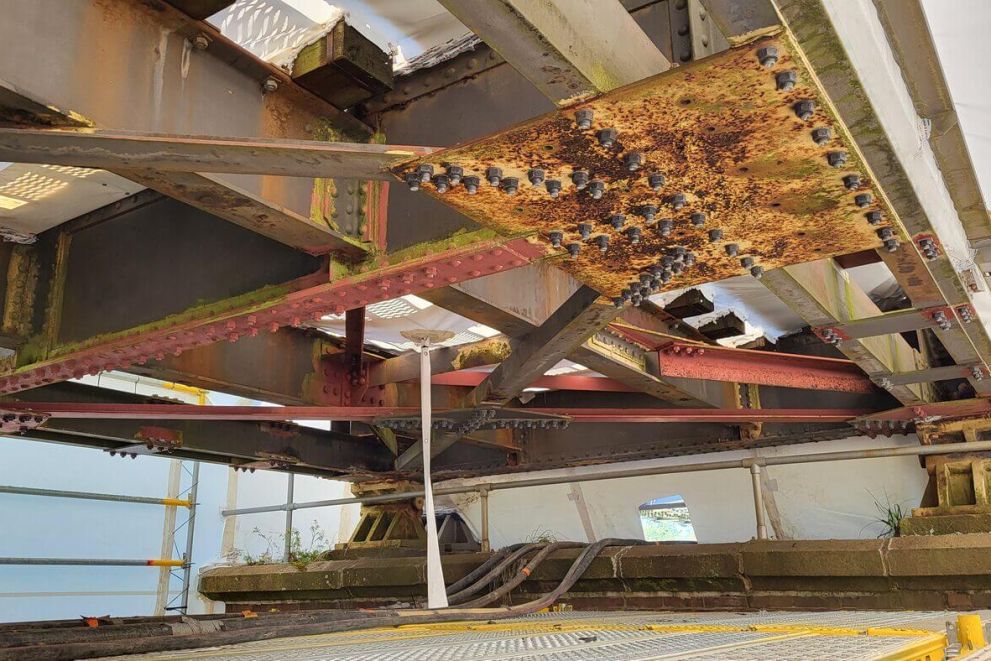 Rostige Stahlträger mit vielen Bolzen ruhen über einem Betonfundament, umgeben von Gerüst und weißer Plane.